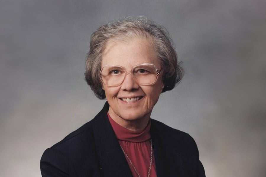 Sister Mary wears a black blazer, burgundy shirt, and square glasses