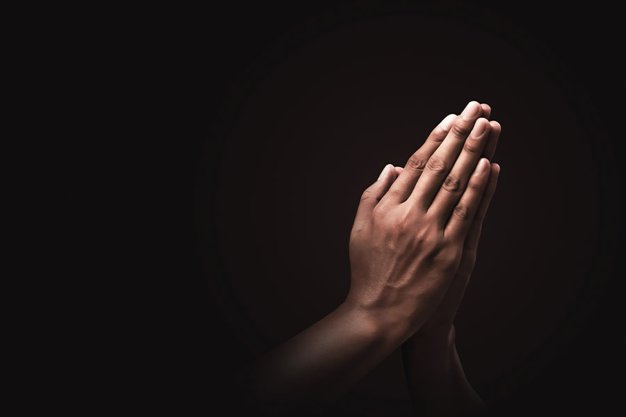A close-up of hands clasped in prayer against a black background.