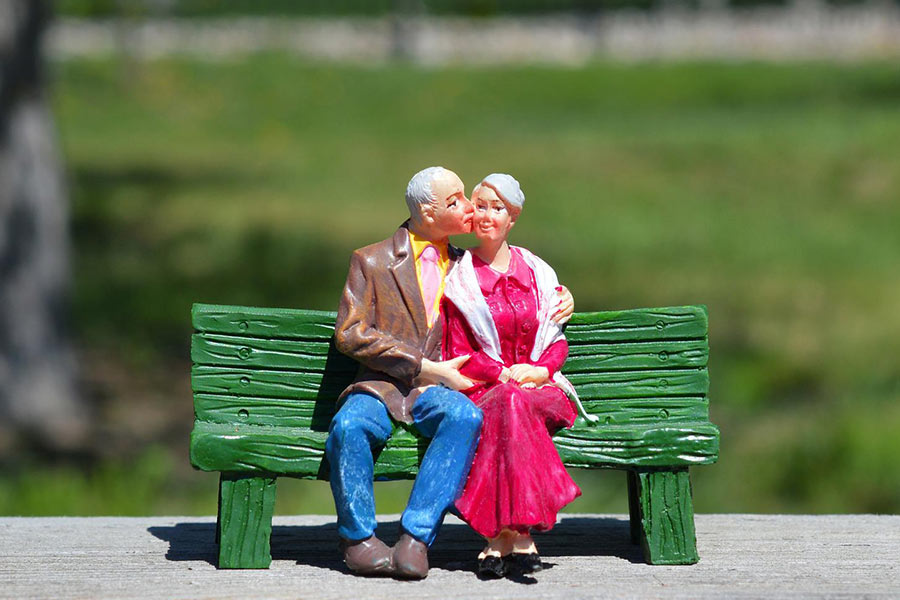 Husband and wife figurines sitting on a park bench.