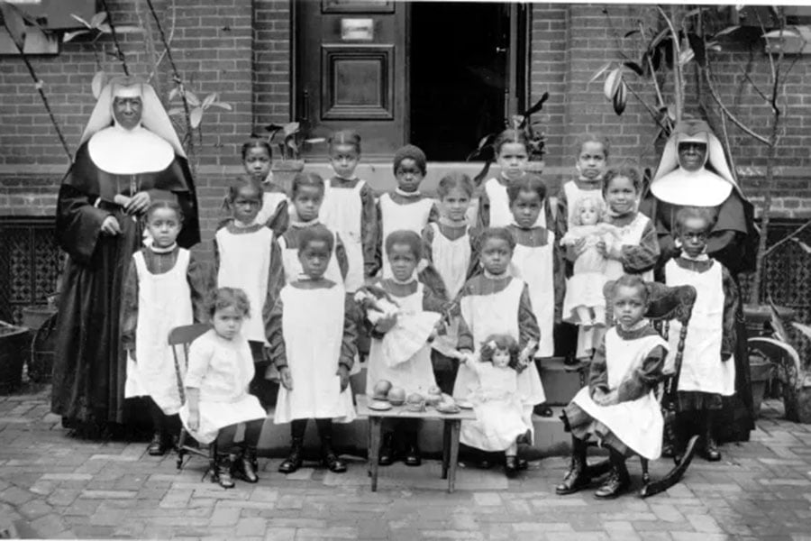 In this black and white picture, eighteen young girls are arranged for a class picture wearing black dresses with white aprons and two sisters border the group on either side.