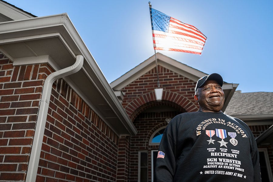 George Webster stands in front of his home with a large American flag waving behind him